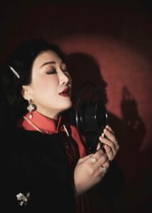 Asian female vocalist singing passionately in a studio environment, holding a microphone.